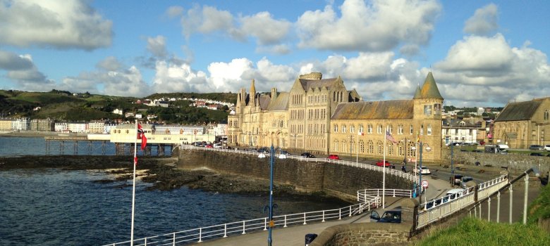 Strandpromenade von Aberystwyth Ein sonniger Tag an der Strandpromenade von Aberystwyth. Im Hintergrund steht das prachtvolle, schlossartige Old College Gebäude aus hellem Stein direkt am Meer unter einem blau-weißen Wolkenhimmel