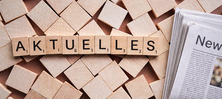 Draufsicht auf quadratische Holzplättchen mit Buchstaben, die das Wort ‚AKTUELLES‘ formen. Die Buchstaben haben kleine Zahlenwerte wie bei einem Scrabble-Spiel. Rechts im Bild ragt der Anschnitt einer Zeitung mit der Aufschrift ‚New‘ ins Bild