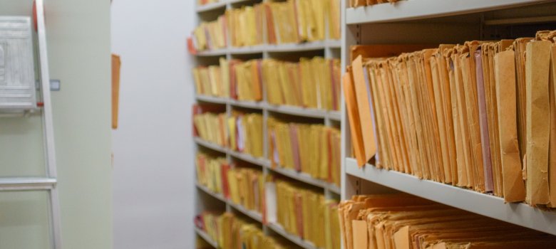 Mit alten Dokumentenbündeln vollgepacktes Regal. Links davon steht eine faltbare Leiter