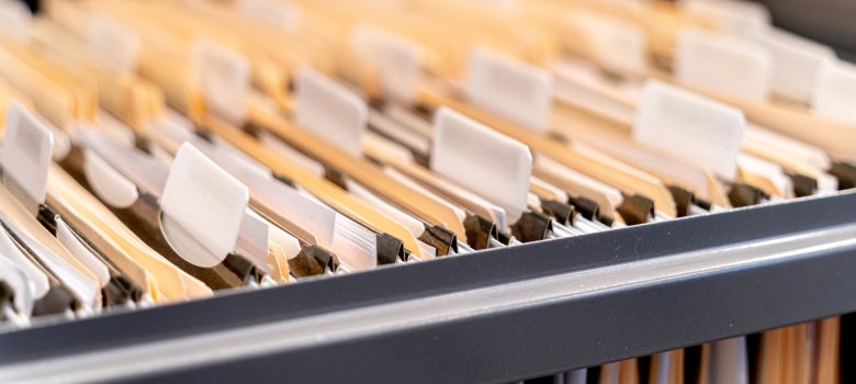Blick von schräg oben in eine geöffnete graue Metallschublade; darin befinden sich zahlreiche beige Hängemappen mit leeren weißen Plastikreitern zur Beschriftung