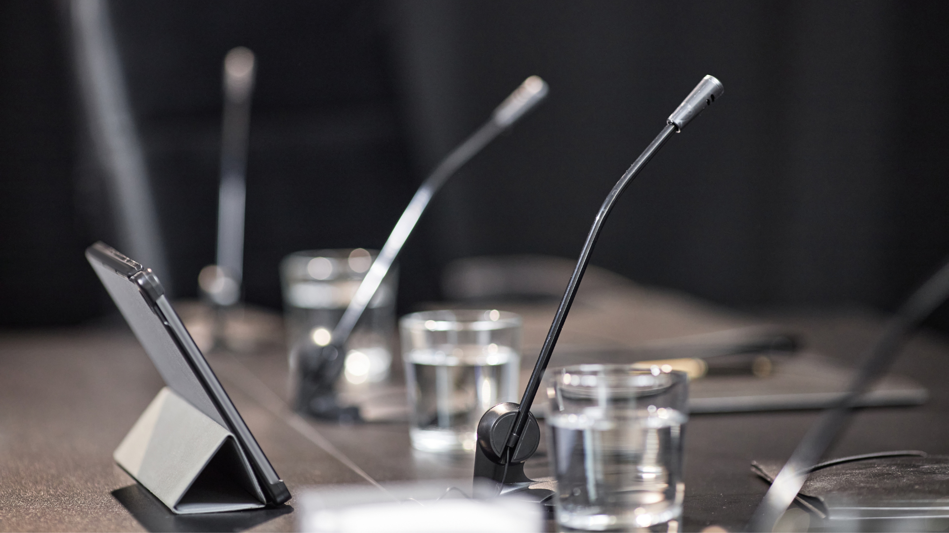 Aktuelles - Gremien - Sitzung Nahaufnahme eines Konferenztisches mit Schwanenhalsmikrofonen, Wassergläsern und einem aufgestellten Tablet