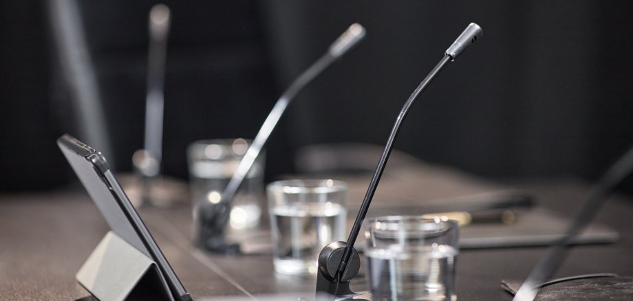 Aktuelles - Gremien - Sitzung Nahaufnahme eines Konferenztisches mit Schwanenhalsmikrofonen, Wassergläsern und einem aufgestellten Tablet