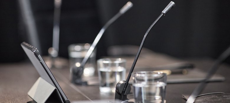 Nahaufnahme eines Konferenztisches mit Schwanenhalsmikrofonen, Wassergläsern und einem aufgestellten Tablet