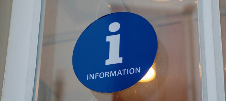 Nahaufnahme eines kreisförmigen blauen Stickers auf Glas. In der Mitte steht ein weißes, kleingeschriebenes ‚i‘, darunter in Versalien das Wort ‚INFORMATION‘. Im Hintergrund sind unscharfe Lichtreflexe einer Innenbeleuchtung zu sehen