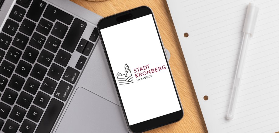 Draufsicht auf einen hölzernen Schreibtisch. In der Mitte liegt ein Smartphone, das auf dem Display das Logo der Stadt Kronberg im Taunus zeigt. Links ist ein Teil einer silbernen Laptop-Tastatur zu sehen, rechts ein weißes liniertes Blatt Papier mit einem weißen Kugelschreiber