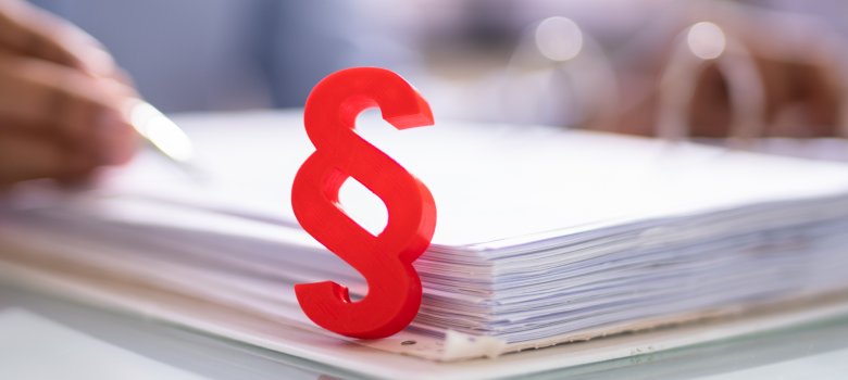 Ein rotes, dreidimensionales Paragraphenzeichen (§) steht im Vordergrund auf einem aufgeschlagenen Aktenordner voller Dokumente. Im unscharfen Hintergrund ist im Anschnitt eine Hand zu sehen, die mit einem Stift auf einem Papier schreibt