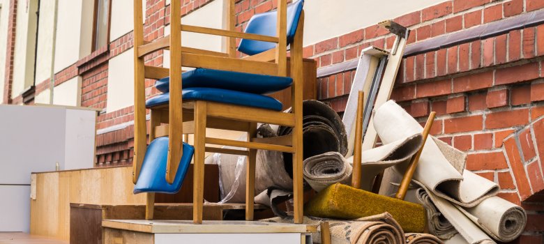 Aktuelles - Sperrmüll 2 Sperrmüll-Ansammlung im Freien: Zwei Holzstühle mit leuchtend blauen Polstern sind auf alten Holzkommoden gestapelt. Daneben liegen mehrere zusammengerollte Teppiche und Polsterreste vor einer roten Backsteinmauer