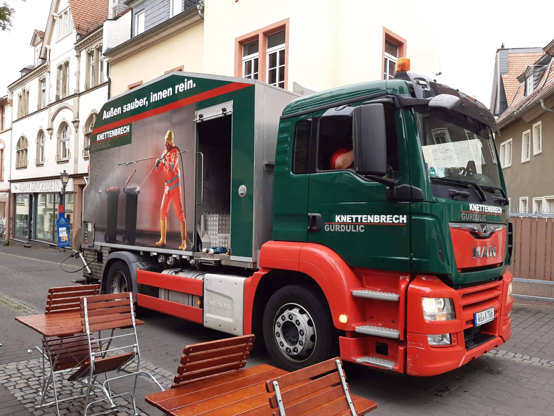 Ein grün-roter Lkw von Knettenbrech & Gurdulic steht in einer Fußgängerzone. Auf dem Fahrzeug zeigt ein großes Werbeplakat die Reinigung einer Mülltonne mit dem Slogan „Außen sauber, innen rein.“.