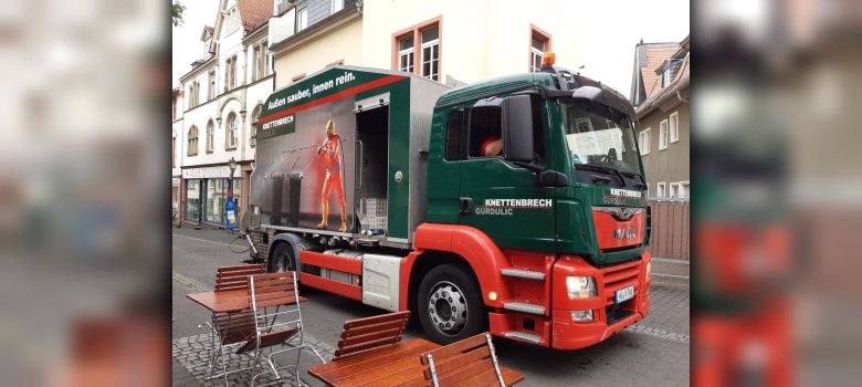 Ein grün-roter Lkw von Knettenbrech & Gurdulic steht in einer Fußgängerzone. Auf dem Fahrzeug zeigt ein großes Werbeplakat die Reinigung einer Mülltonne mit dem Slogan „Außen sauber, innen rein.“.