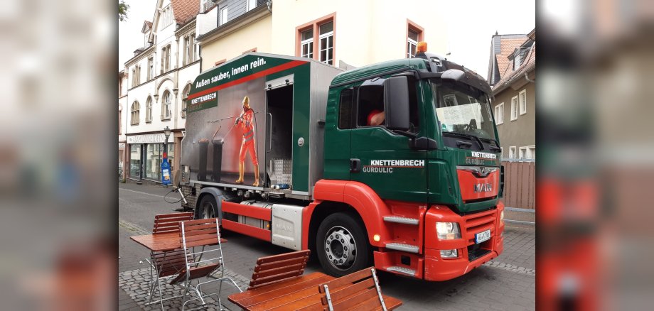 Ein grün-roter Lkw von Knettenbrech & Gurdulic steht in einer Fußgängerzone. Auf dem Fahrzeug zeigt ein großes Werbeplakat die Reinigung einer Mülltonne mit dem Slogan „Außen sauber, innen rein.“.