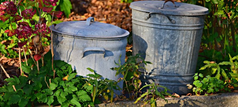 Zwei klassische, geschlossene Mülltonnen aus verzinktem Metall stehen nebeneinander in einem Gartenbeet hinter einer niedrigen Betonmauer, umgeben von grünen Pflanzen und einer blühenden Hortensie.