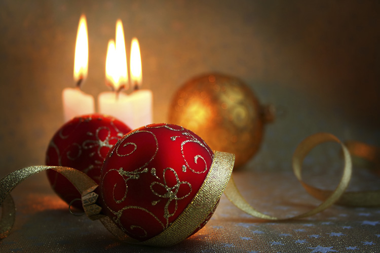 Ein stimmungsvolles Stillleben mit zwei roten, gold verzierten Weihnachtskugeln im Vordergrund und drei brennenden Kerzen im unscharfen Hintergrund