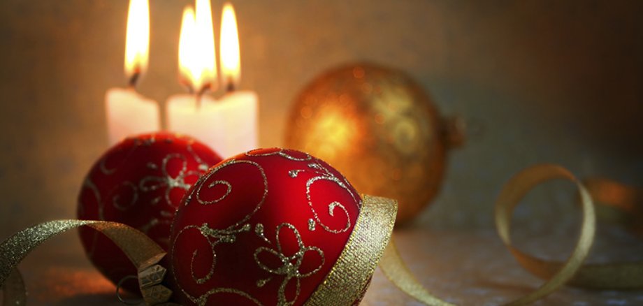 Ein stimmungsvolles Stillleben mit zwei roten, gold verzierten Weihnachtskugeln im Vordergrund und drei brennenden Kerzen im unscharfen Hintergrund