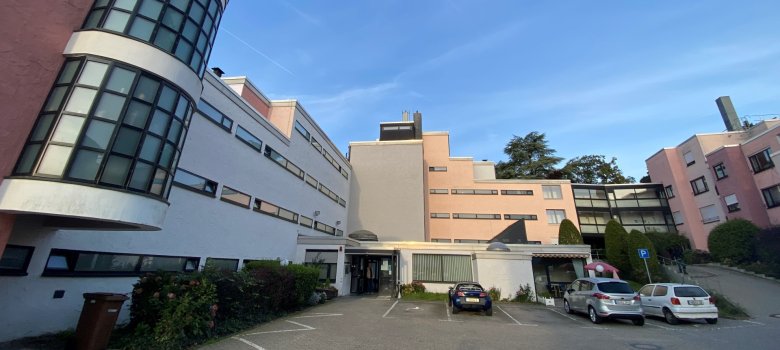 Blick auf einen Gebäudeteil der Siedlung Roter Hang in Kronberg. Zu sehen ist ein mehrstöckiges, weißes Gebäude mit markantem, halbrundem Glasturm und schmalen Fensterbändern vor einem Parkplatz mit Autos