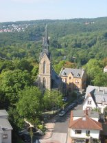Eine Aufnahme aus erhöhter Position auf ein Stadtviertel bei Tageslicht. Im Zentrum steht eine Kirche aus hellem Naturstein mit einem sehr steilen, dunklen Turmhelm. Die Kirche ist von hohen grünen Bäumen umgeben. Rechts daneben verläuft eine Straße mit parkenden Autos und mehrstöckigen Wohnhäusern mit Ziegeldächern. Der Hintergrund wird von einem dichten, weiten Waldgebiet dominiert, das sich über sanfte Hügel erstreckt. In der Ferne sind am Hang weitere helle Häuser einer Siedlung erkennbar