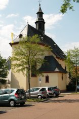 Die Außenansicht einer einschiffigen Kirche bei Tageslicht. Das Gebäude hat hellgelb verputzte Wände mit rötlichen Sandsteinfassungen an den hohen Rundbogenfenstern. Das steile, dunkle Schieferdach trägt einen achteckigen Dachreiter mit Turmuhr und spitzer Haube. An der linken Dachecke weht eine gelb-weiße Fahne. Im Vordergrund stehen mehrere Autos auf einem gepflasterten Parkplatz, der durch junge Bäume von der Kirche getrennt ist. Der Himmel ist leicht bewölkt