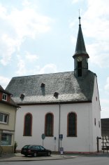 Die Außenaufnahme zeigt eine schlichte, weiß verputzte Kirche mit drei hohen Rundbogenfenstern an der Längsseite. Das massive, steile Dach ist mit dunklem Schiefer gedeckt und besitzt drei kleine Gauben. Auf der rechten Giebelseite thront ein schmaler, achteckiger Turm mit einer Turmuhr und einer sehr spitzen Schieferhaube. Im Vordergrund parkt ein schwarzer Kombi vor dem Gebäude an einer Straße. Der Himmel ist hell mit vereinzelten weißen Wolken