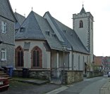 Die Außenaufnahme zeigt eine mittelalterliche Kirche in einer engen städtischen Gasse. Das Gebäude hat hell verputzte Wände mit rötlichen Sandstein-Einfassungen an den spitzbogigen Maßwerkfenstern. Ein sehr steiles, verwinkeltes Dach aus dunklem Schiefer bedeckt das Kirchenschiff. Rechts ragt ein massiver, quadratischer Turm mit einer zinnenbewehrten Plattform und einem kleinen spitzen Dachhelm empor. Eine niedrige Steinmauer mit einem schmiedeeisernen Tor trennt das Kirchengelände von der asphaltierten Straße. Links im Vordergrund ist die schieferverkleidete Fassade eines Nachbarhauses zu sehen