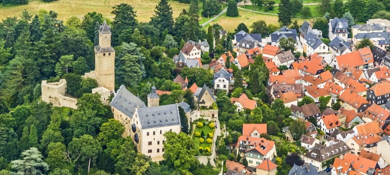 Ein Panoramafoto aus der Vogelperspektive auf die Burg Kronberg. Links im Bild steht der hohe, schlanke Freiturm aus hellem Stein neben massiven Burgmauern. Das Hauptgebäude der Burg mit seinem dunklen Schieferdach und kleinen Gauben liegt zentral in einer grünen Parkanlage mit einem geometrisch angelegten Garten. Rechts schließt sich die dichte Bebauung der Kronberger Altstadt mit ihren charakteristischen orange-roten Ziegeldächern und hellen Fassaden an. Der gesamte Komplex ist in eine weitläufige Wald- und Wiesenlandschaft eingebettet