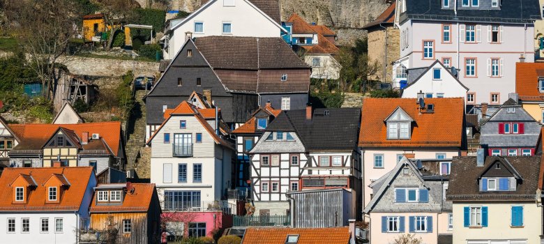 Eine dichte Ansammlung von historischen Häusern an einem steilen Hang. Im Vordergrund stehen verschiedene Fachwerkhäuser mit weißen Fassaden, dunklem Gebälk und orangefarbenen Ziegeldächern. Darüber ziehen sich weitere Wohnhäuser mit hellen Putzfassaden und dunklen Schieferdächern den Hang hinauf. Den oberen Abschluss bildet eine massive, mittelalterliche Steinmauer der Burg Kronberg, hinter der ein runder Wehrturm mit Fachwerkobergeschoss (der Pulverturm) und die Mauern des Palas aufragen