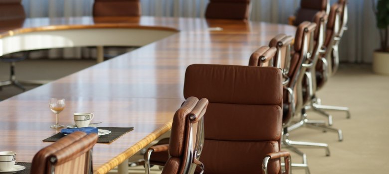 Nahaufnahme eines Konferenzbereiches mit Fokus auf die Bestuhlung. Im Vordergrund stehen mehrere Design-Bürostühle aus braunem Leder mit Chromgestell an einem glänzenden, ovalen Echtholztisch. Auf dem Tisch stehen vereinzelt Kaffeetassen und ein Weinglas auf dunklen Platzsets. Im Hintergrund setzt sich die Sitzreihe an dem geschwungenen Tisch fort, davor hängen bodentiefe, helle Vorhänge, die das Tageslicht sanft filtern