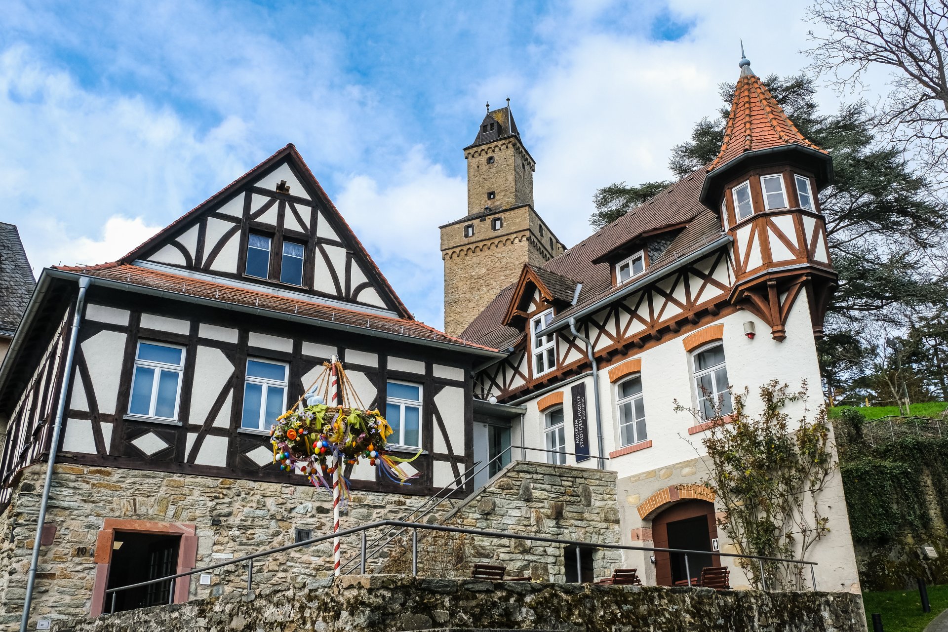 Ein Ensemble aus historischen Gebäuden, darunter ein Fachwerkhaus auf einem Steinsockel und ein weißes Haus mit einem spitzen Ecktürmchen, hinter denen ein massiver viereckiger Steinturm aufragt
