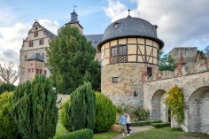 Ein runder, historischer Festungsturm aus Naturstein mit einem Fachwerkobergeschoss und Schieferdach steht in einer Gartenanlage, während im Vordergrund ein Paar durch einen Steinbogen geht.