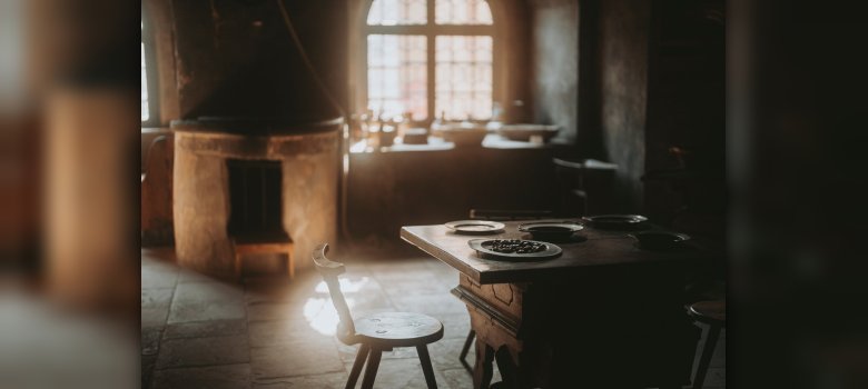 Eine historische Burgküche mit einem rustikalen Holztisch und Stühlen im Vordergrund, auf dem metallene Teller stehen, während im Hintergrund ein gemauerter Ofen und ein großes Fenster im warmen Gegenlicht zu sehen sind.