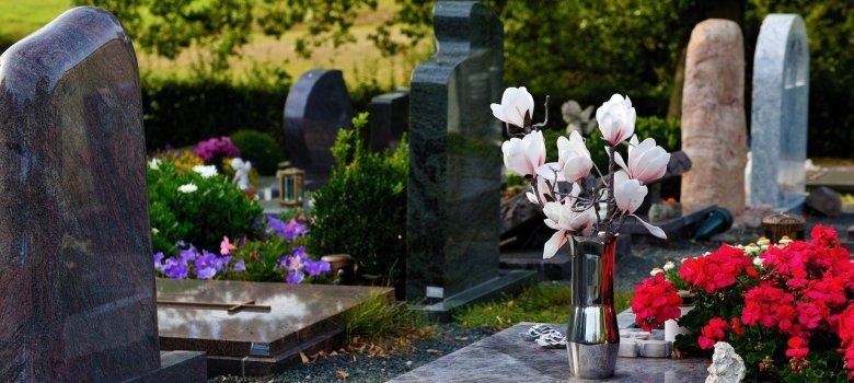 Nahaufnahme einer Grabanlage mit mehreren Grabsteinen in Grau- und Schwarztönen. Im Vordergrund steht eine Edelstahlvase mit weißen Magnolienblüten auf einer polierten Steinplatte neben leuchtend roten Blumen