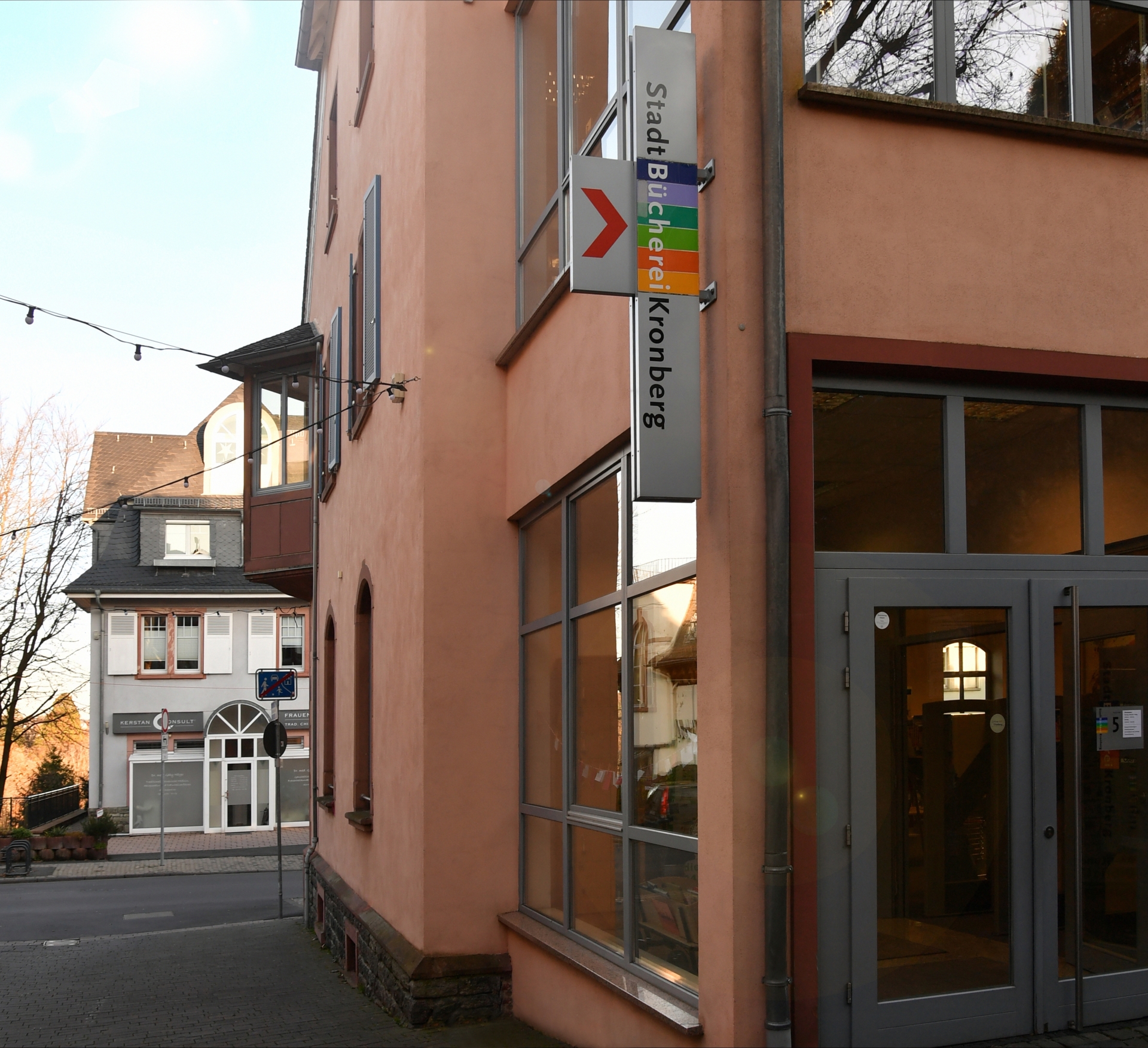 Die Außenansicht des Eingangsbereichs der StadtBücherei Kronberg mit einem vertikalen, farbigen Hinweisschild an der lachsfarbenen Fassade.