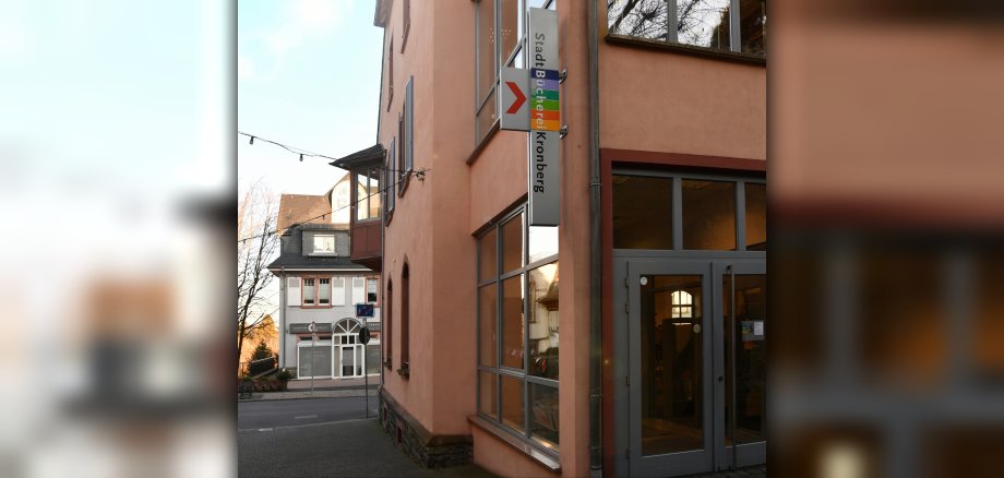 Die Außenansicht des Eingangsbereichs der StadtBücherei Kronberg mit einem vertikalen, farbigen Hinweisschild an der lachsfarbenen Fassade.