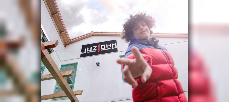 Ein Jugendlicher mit Locken und roter Daunenjacke posiert gut gelaunt im Vordergrund vor dem Gebäude und dem Logo des Jugendzentrums „Juze DHO“.