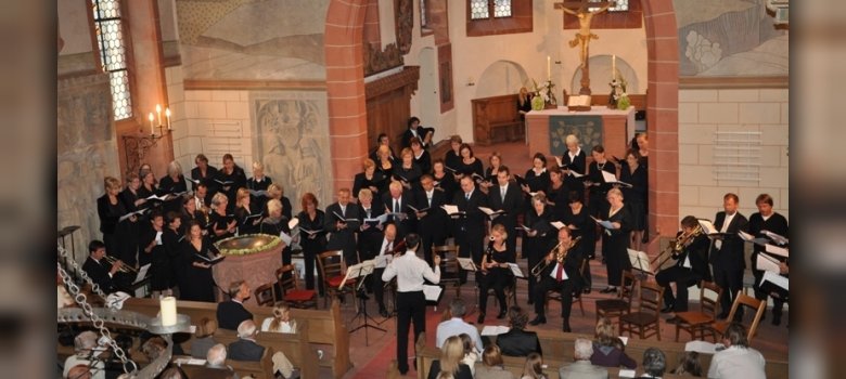 Ein Chor und ein kleines Orchester musizieren unter der Leitung eines Dirigenten im Altarraum einer historischen, kunstvoll ausgemalten Kirche