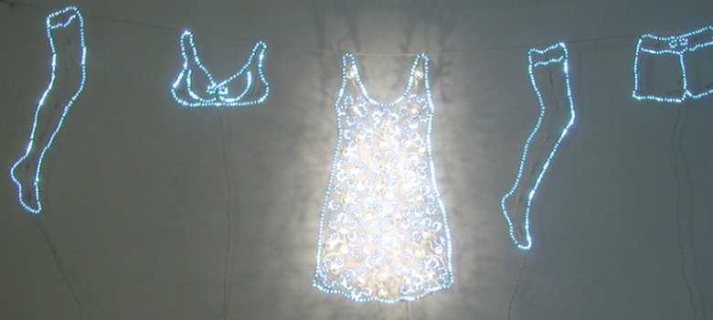 Eine leuchtende Wäscheleine-Installation an einer Wand mit aus LED-Lichterketten geformten Kleidungsstücken wie Kleid, BH, Shorts und Beinen.