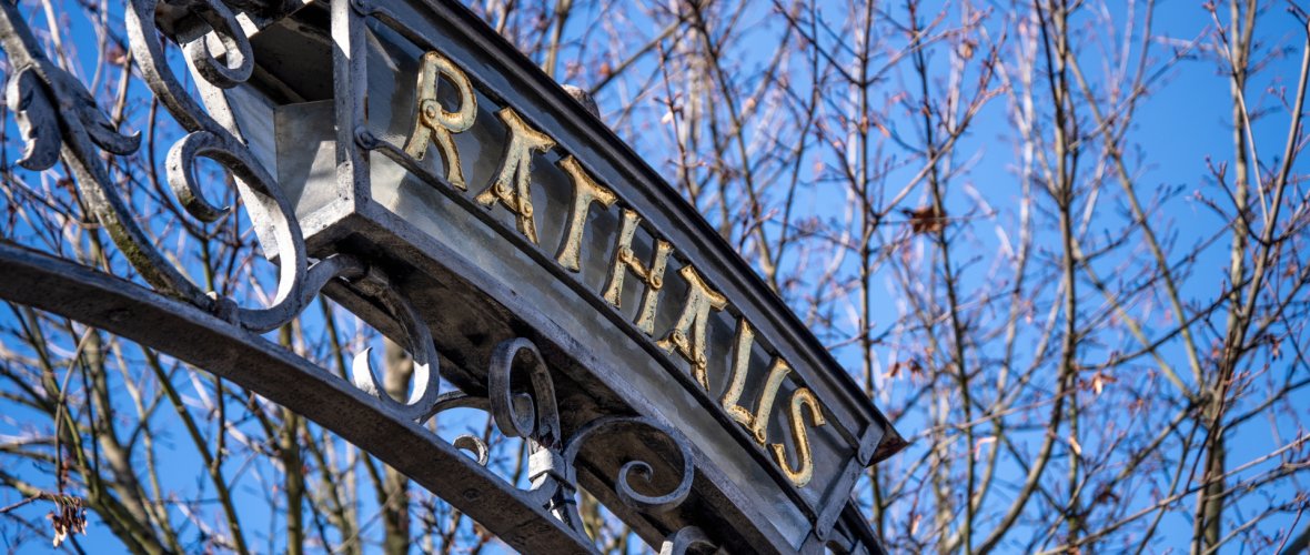 Die Perspektive von schräg unten auf den Scheitelpunkt eines kunstvoll geschmiedeten Metallbogens. In einem rechteckigen, leicht geschwungenen Metallrahmen prangen goldene Großbuchstaben, die das Wort ‚RATHAUS‘ bilden. Der dunkelgraue Bogen ist mit filigranen Schnörkeln und Blattmotiven verziert. Im Hintergrund sind kahle Baumzweige und ein klarer, tiefblauer Winter- oder Vorfrühlingshimmel zu sehen. Das Sonnenlicht lässt die goldenen Buchstaben hell erstrahlen
