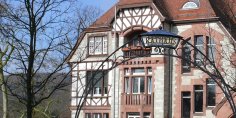 Ein filigran geschmiedeter Torbogen mit dem goldenen Schriftzug „RATHAUS“ steht im Vordergrund vor der historischen Fachwerk- und Natursteinfassade des Kronberger Rathauses unter blauem Himmel.