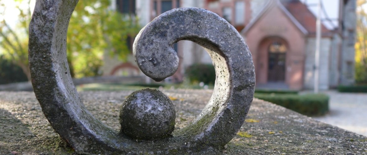 Eine extreme Nahaufnahme eines geschmiedeten oder gegossenen Metallornaments in Form einer Schnecke oder Spirale. Das dunkle, wettergegerbte Metall weist eine raue, punktierte Oberfläche auf und ruht auf einer flachen, hellgrauen Steinmauer. Im Zentrum der Spirale liegt eine kleine Metallkugel. Der Hintergrund ist in weicher Unschärfe gehalten und lässt die rötliche Sandsteinfassade und das Portal der Villa Bonn bei hellem Tageslicht erkennen