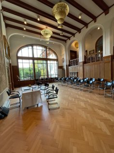 Der holzgetäfelte Festsaal des Kronberger Rathauses mit edlem Fischgrätparkett, hohen Deckenbögen, kunstvollen Kronleuchtern und einer Bestuhlung für eine offizielle Sitzung oder Trauung vor einer großen Fensterfront.