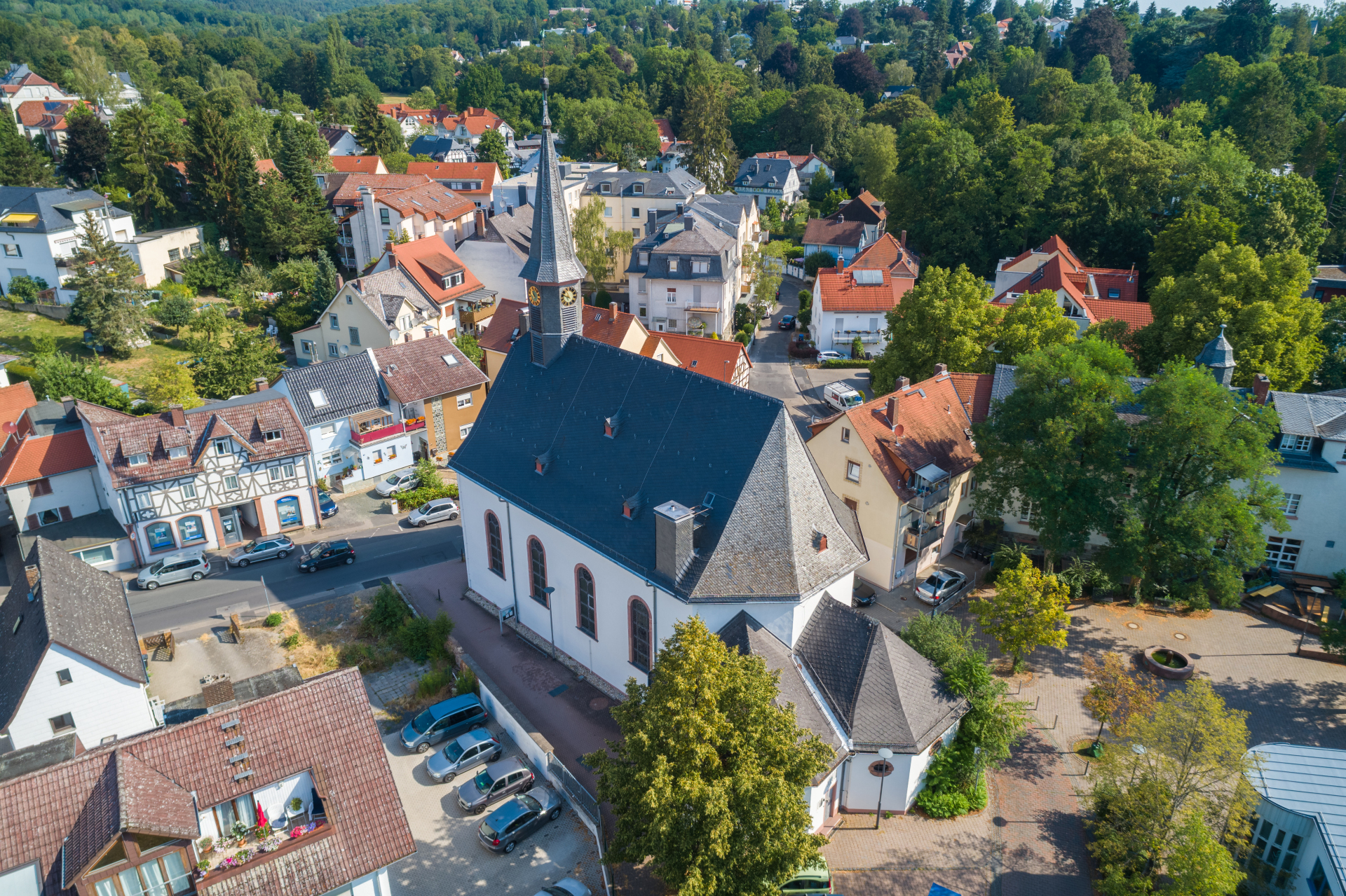Eine Luftaufnahme einer historischen Kirche mit spitzem Kirchturm, umgeben von Wohnhäusern, Straßen und dichten grünen Bäumen.