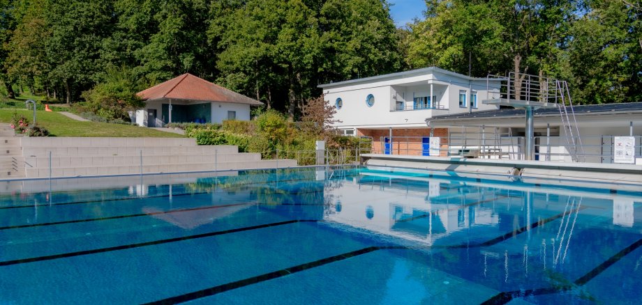Blick über das blaue, spiegelglatte Sportbecken im Kronberger Waldschwimmbad mit den Startbahnen, dem Sprungturm und den Funktionsgebäuden vor einer dichten Waldkulisse.