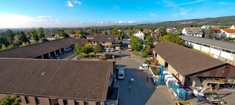 Eine Luftaufnahme eines weitläufigen Betriebshofs der Stadtwerke Kronberg mit mehreren Hallen, Fahrzeugen und Besuchern unter blauem Himmel.