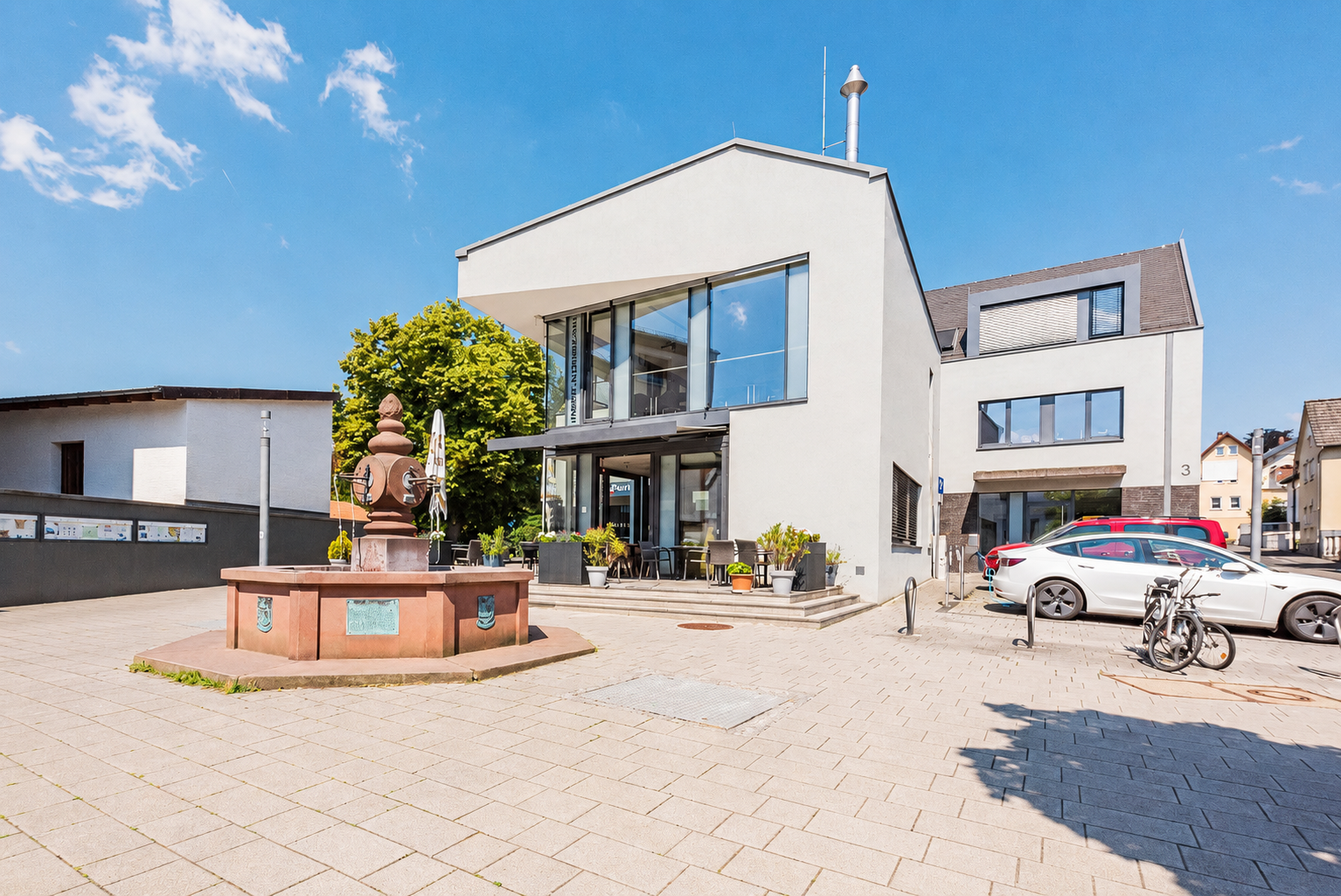 Ein gepflasterter Platz mit einem historischen Brunnen vor einem modernen, weißen Gebäude mit dem Restaurant „Punjab Tandoori“ unter blauem Himmel.