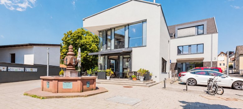 Ein gepflasterter Platz mit einem historischen Brunnen vor einem modernen, weißen Gebäude mit dem Restaurant „Punjab Tandoori“ unter blauem Himmel.