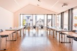 Ein großer, heller Veranstaltungsraum mit dunklem Parkettboden, einer hufeisenförmigen Tischordnung mit schwarzen Stühlen und einer bodentiefen Fensterfront mit Ausblick auf Hausdächer