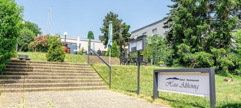 Eine Außenansicht des Hotels und Restaurants „Haus Altkönig“ mit einer breiten Waschbetontreppe, die zur Außenterrasse und zum Gebäude führt, sowie einem Schild im Vordergrund.