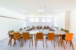 Ein heller, funktionaler Konferenzraum mit weißen Wänden, quadratischen Deckenleuchten und in U-Form angeordneten Holztischen samt passenden Holzstühlen mit grünen Sitzpolstern.