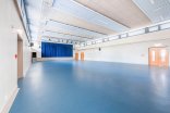 Ein weitläufiger, moderner Veranstaltungssaal mit blauem Linoleumboden, hellen Wänden, einer hohen Decke mit Akustikpaneelen und einer Bühne mit geschlossenem, blauem Vorhang am Ende des Raumes