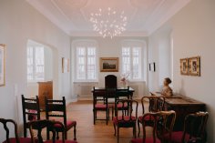 Ein elegant eingerichtetes Trauzimmer oder historischer Raum mit Stuckdecke, einem zentralen Schreibtisch, antiken Holzmöbeln, roten Polsterstühlen und kunstvollen Gemälden an den hellen Wänden.
