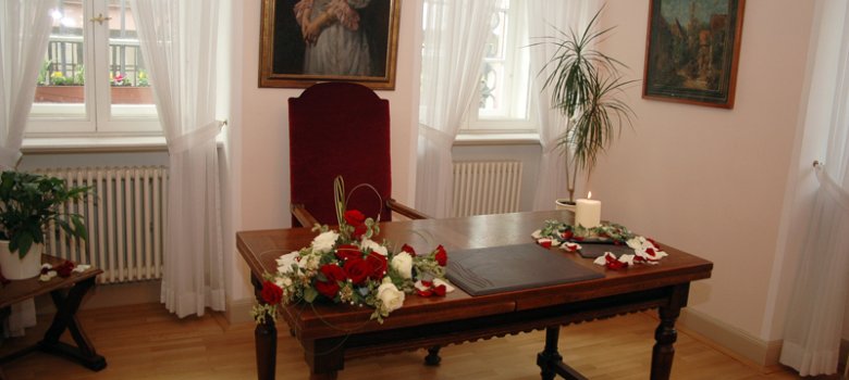 Innenansicht eines hellen Zimmers mit zwei Fenstern und weißen Vorhängen. Im Zentrum steht ein massiver Holztisch mit einem Gesteck aus Rosen und einer weißen Kerze. Dahinter steht ein hoher, roter Samtstuhl vor einem klassischen Ölgemälde