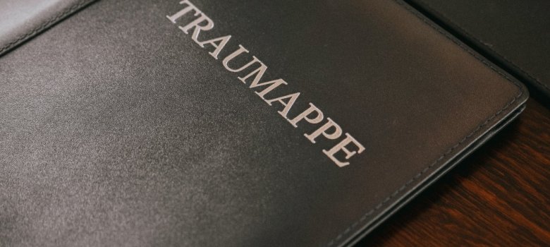 Draufsicht auf eine dunkelgraue oder schwarze Mappe in Lederoptik. In der Mitte ist schräg das Wort ‚TRAUMAPPE‘ in silberner Serifenschrift eingeprägt. Die Mappe liegt auf einem dunkelbraunen Holztisch